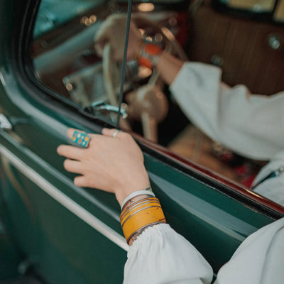 Wearing the Frankie - Dodge pickup adjustable bracelet by Joyride Jewelry, a person rests their hand on the open window of a vintage green Ford car, with another hand on the steering wheel showcasing the classic car’s original paint.