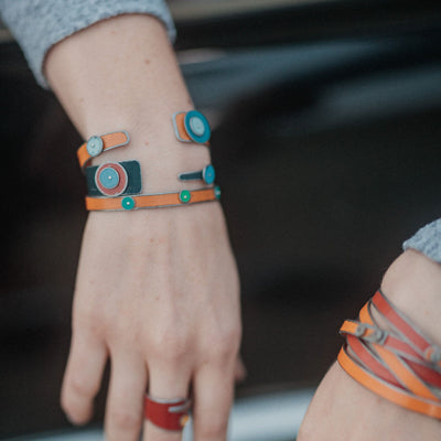 Two hands wearing Billie - Chevy, Oldsmobile, Plymouth wrap bracelets with circular and rectangular blue, green, and orange accents rest on a classic Oldsmobile hood. One hand also features a red ring; the background is blurred.