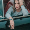 A light brown-haired woman sits in a car with a burgundy interior, gazing thoughtfully at the camera. She wears a gray long-sleeve top and Billie - Chevy & Ford bracelets, resting her arm on the window.