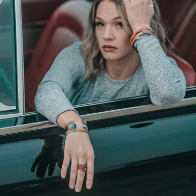 A light brown-haired woman sits in a car with a burgundy interior, gazing thoughtfully at the camera. She wears a gray long-sleeve top and Billie - Chevy & Ford bracelets, resting her arm on the window.