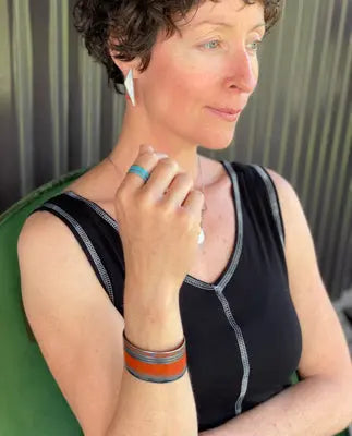 A woman with short curly hair sits thoughtfully in a green chair, wearing a sleeveless black top and the Samson - Ford Falcon wireless earbuds, along with an adjustable turquoise ring and a brown bracelet.