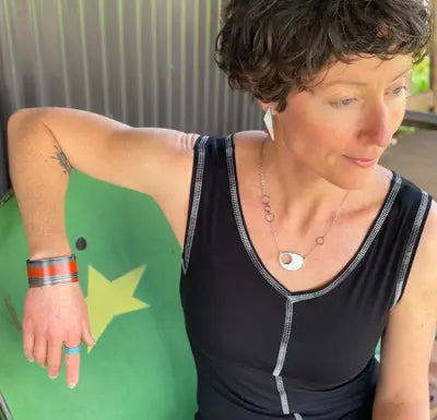 A woman with short curly hair sits. She wears a sleeveless black top,  jewelry by Joyride, an adjustable ring, and colorful bracelets, looking down to her right with a relaxed expression.